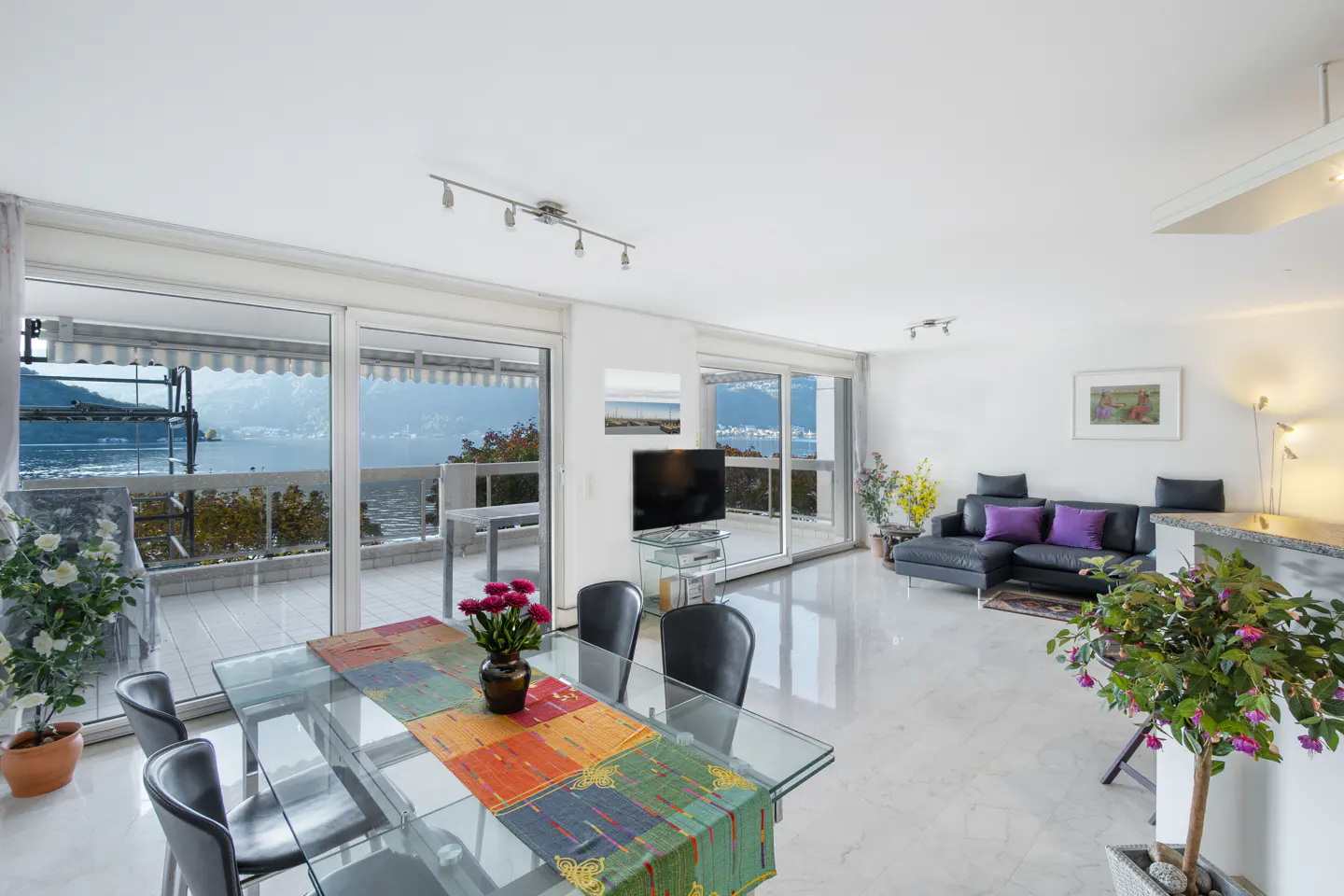 Bright, modern apartment with a glass dining table, black leather sofa, and a balcony overlooking a lake.