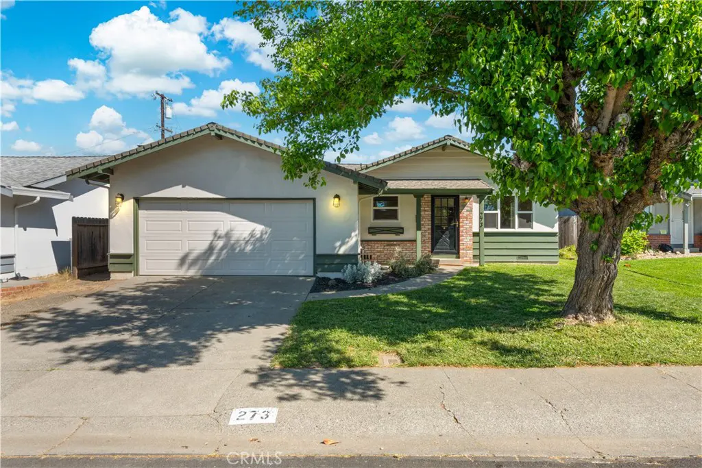A single-story house with a white garage door, green trim, and a large tree in the front yard.