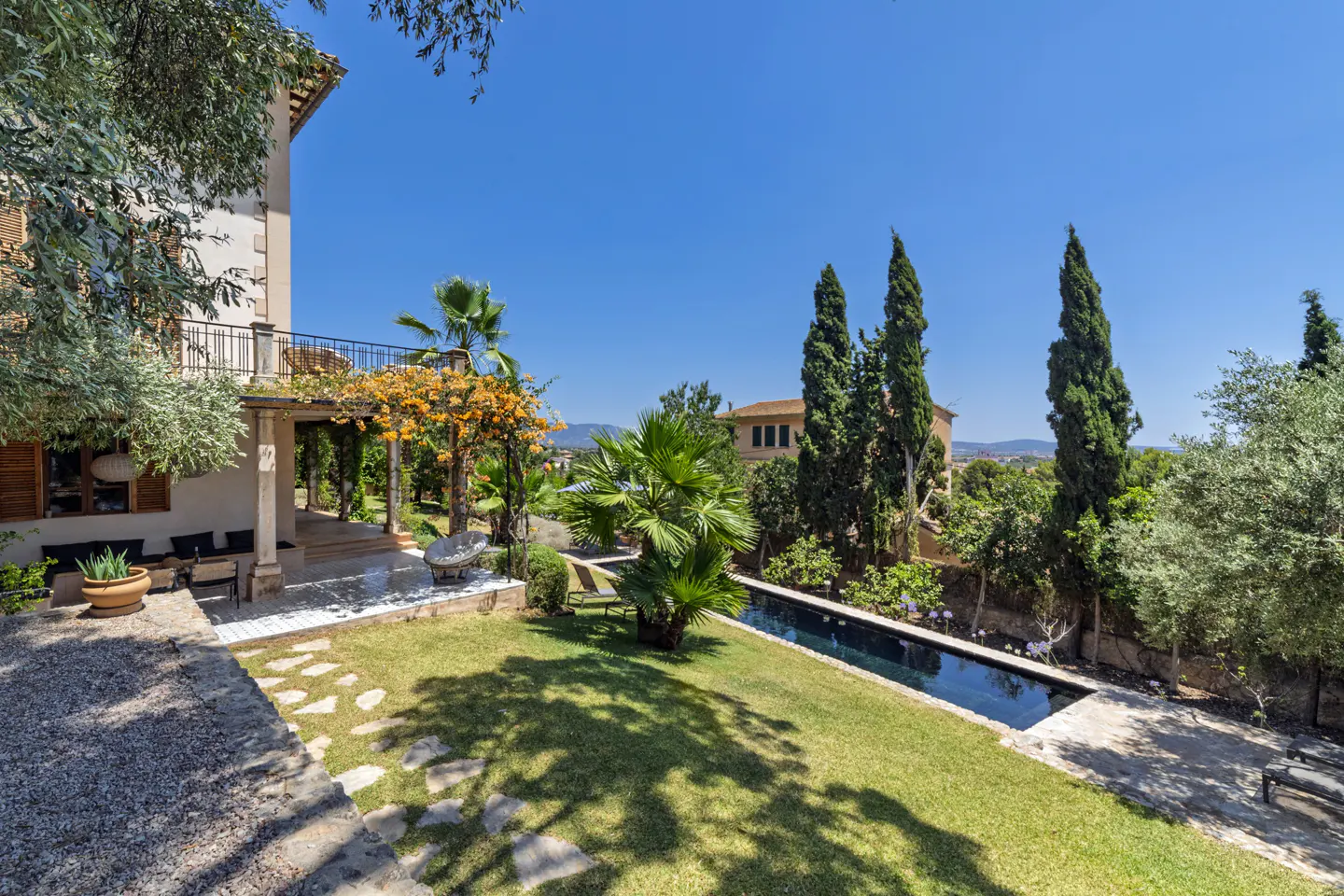 A sunny backyard with a long, narrow pool, green lawn, trees, and a stone patio with orange flowers.