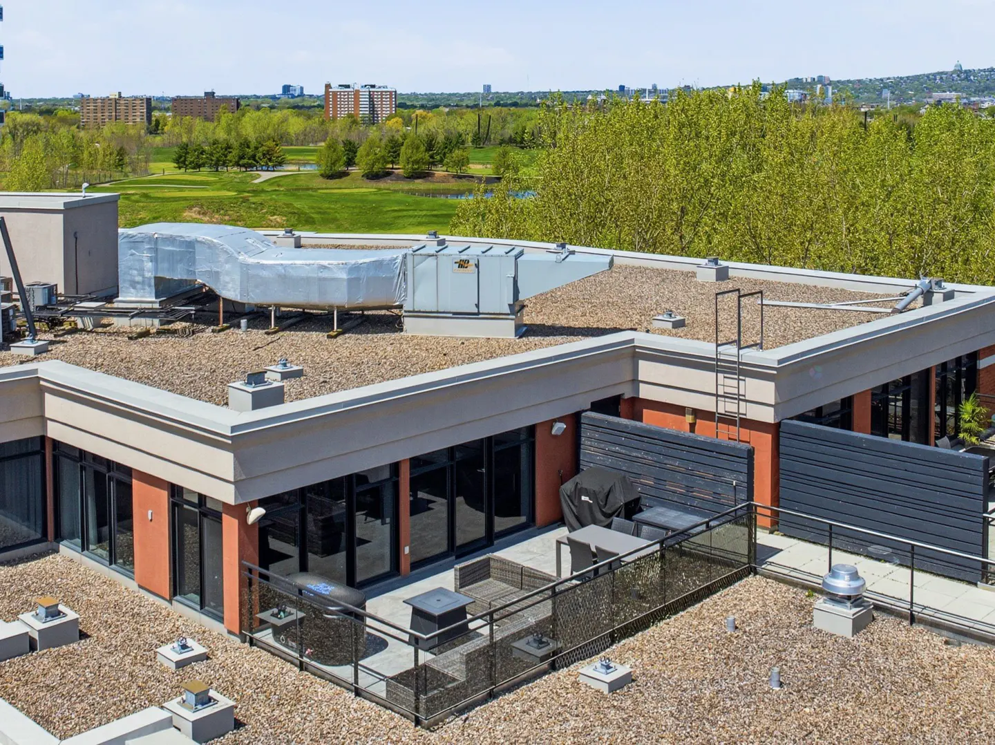 Rooftop view of a modern building with a patio, grill, and outdoor furniture. Green trees and buildings are visible in the background.