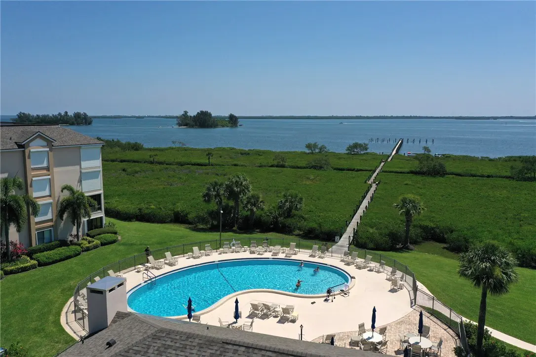 Aerial view of a kidney-shaped pool with people swimming, surrounded by lounge chairs, a building, and a long pier leading to the ocean.