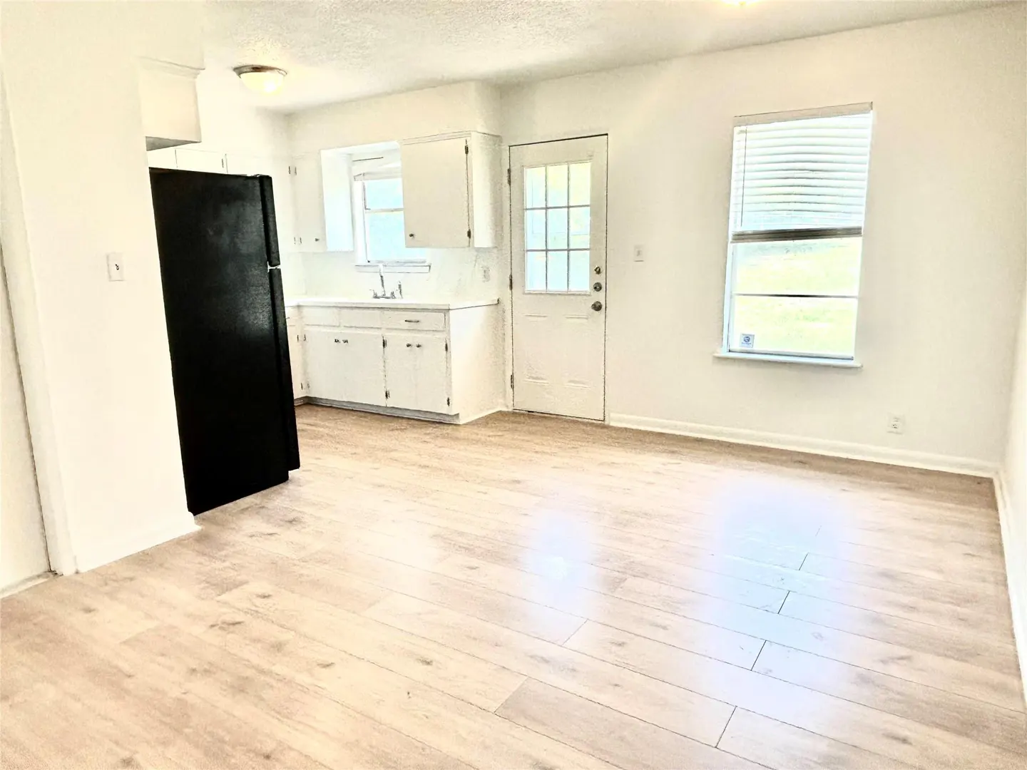 Bright, empty room with light wood floors, white walls, and a black refrigerator. White cabinets and a door are visible in the background.