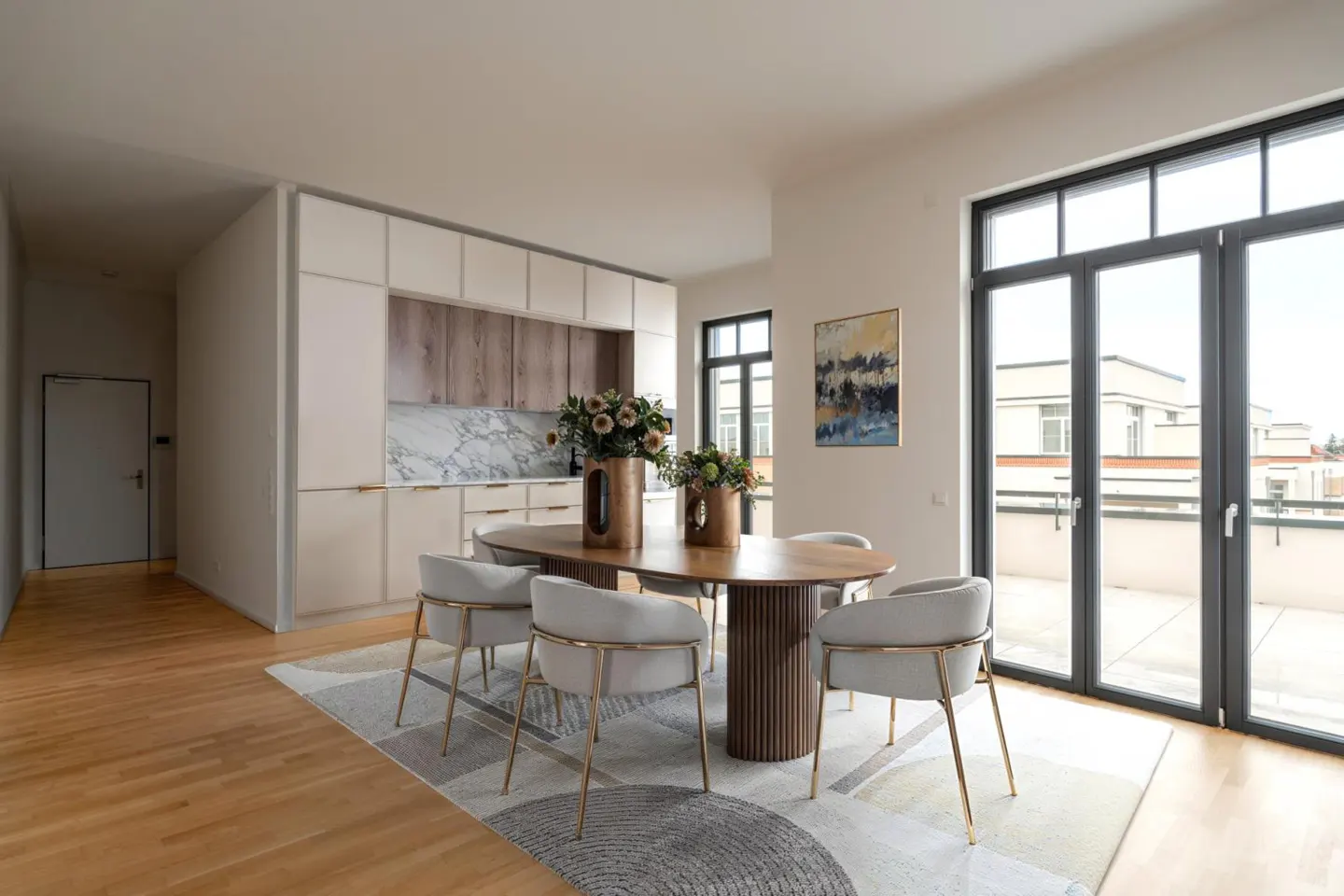 Bright, modern dining area with an oval wood table, six gray chairs, and a patterned rug. Kitchen cabinets and a balcony are visible.