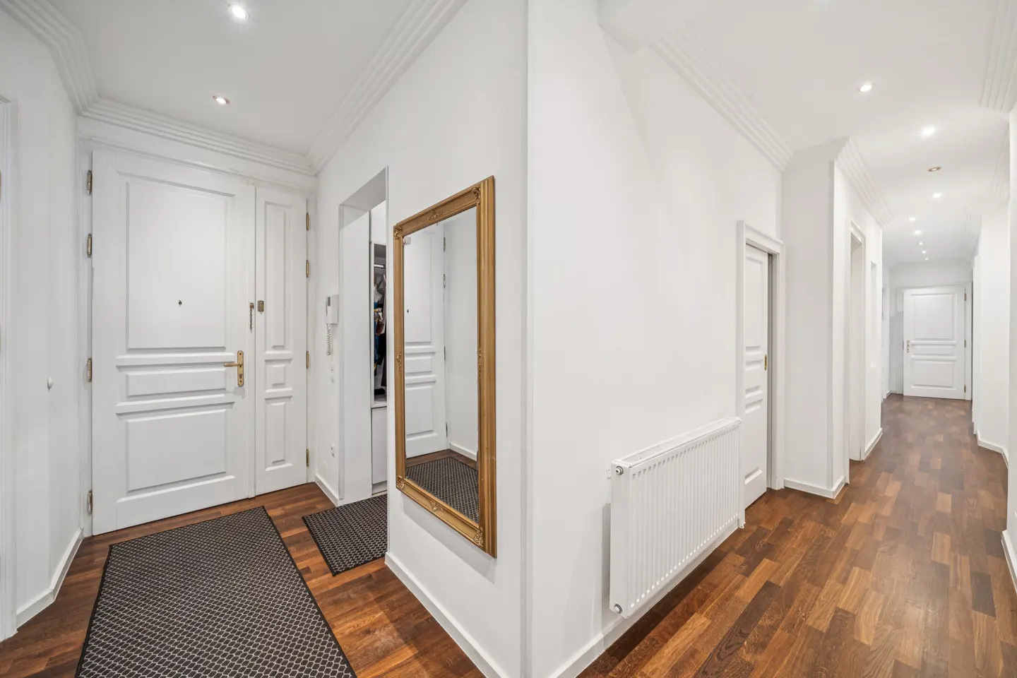Bright white hallway with dark wood floors, a gold-framed mirror, a white radiator, and a white front door.
