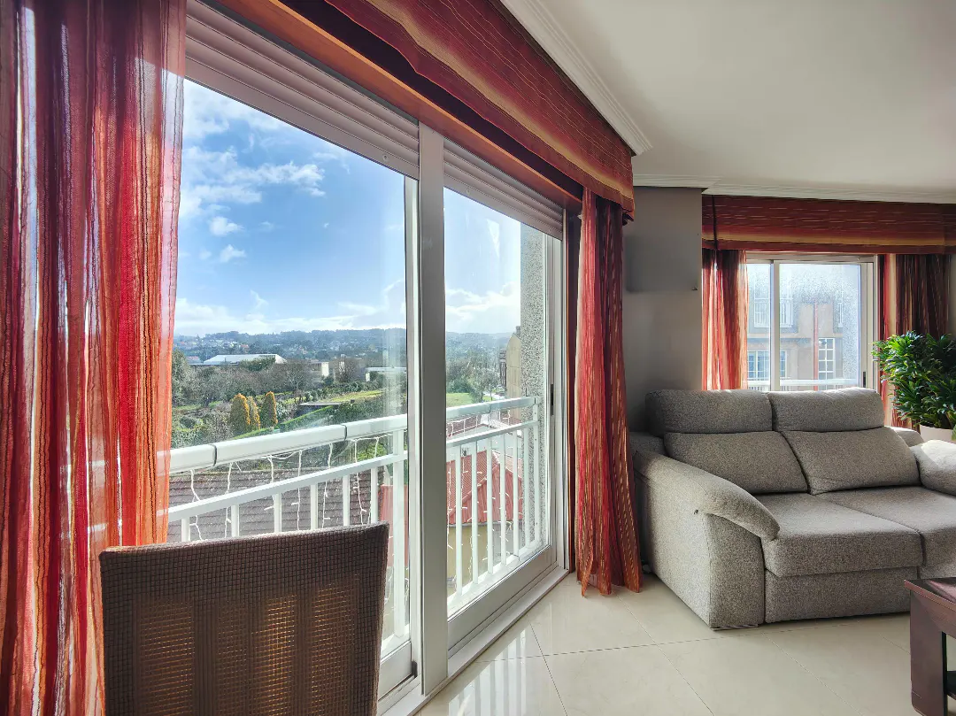 Living room with a grey sofa, red curtains, and a large sliding glass door opening to a balcony with a scenic outdoor view.