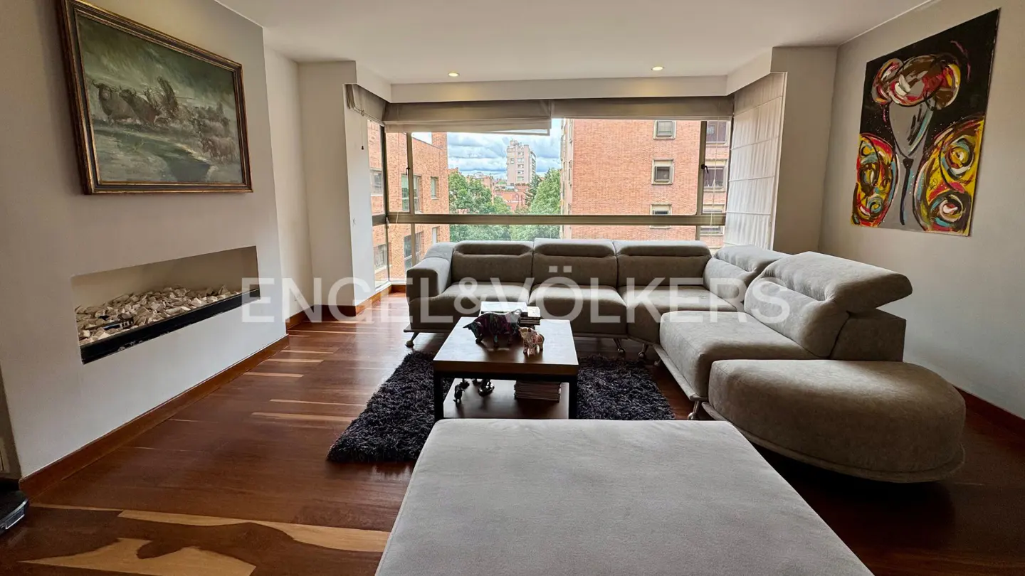 Modern living room with a large grey sectional sofa, wooden floors, a fireplace, and art on the walls facing a city view.