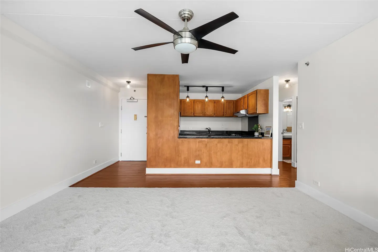 Open-concept living area with white carpet, a ceiling fan, and a kitchen featuring wood cabinets and a breakfast bar.