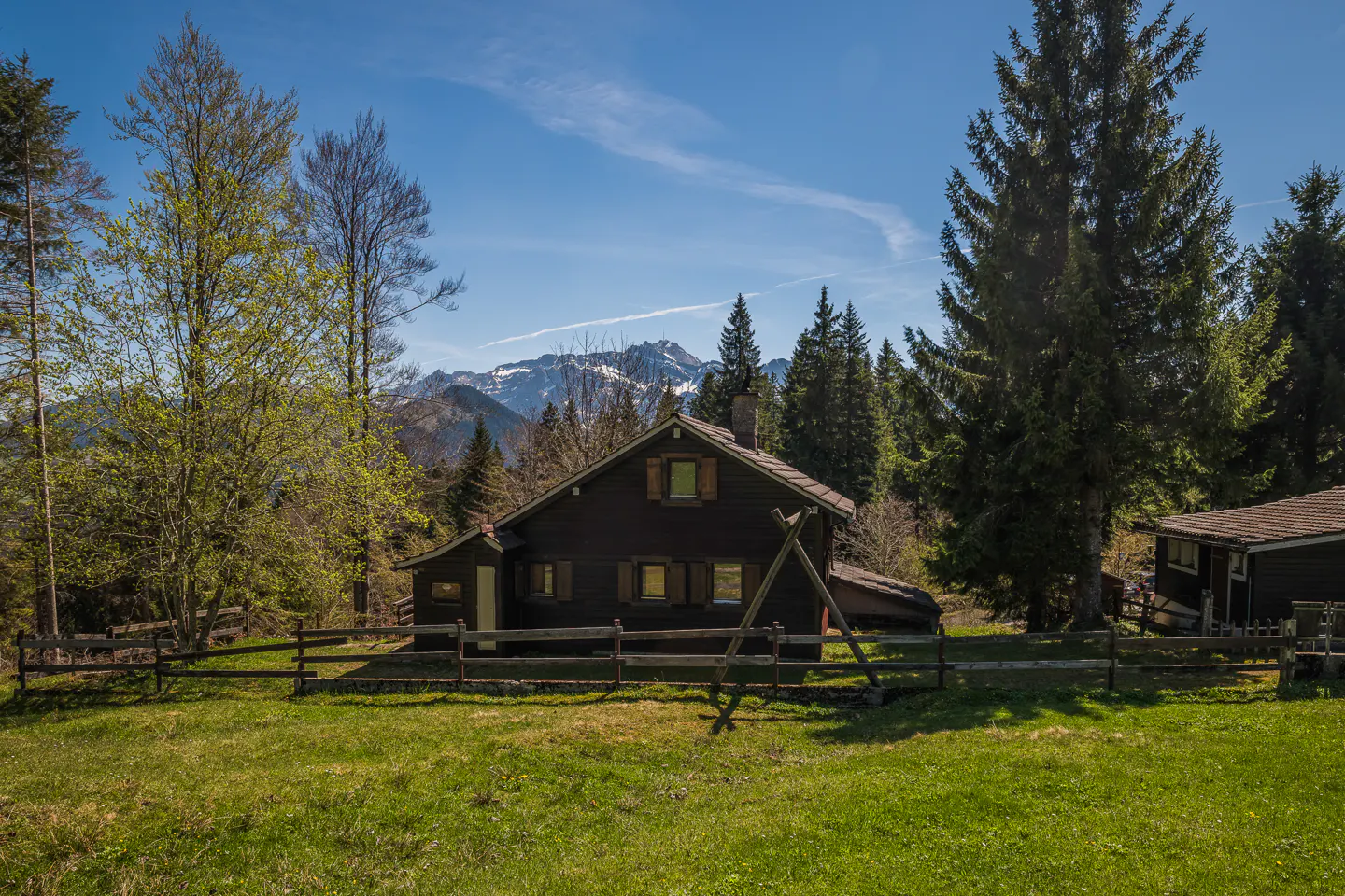 Dark brown cabin with a wooden fence on a green lawn, surrounded by trees. Snow-capped mountains are visible in the background under a blue sky.