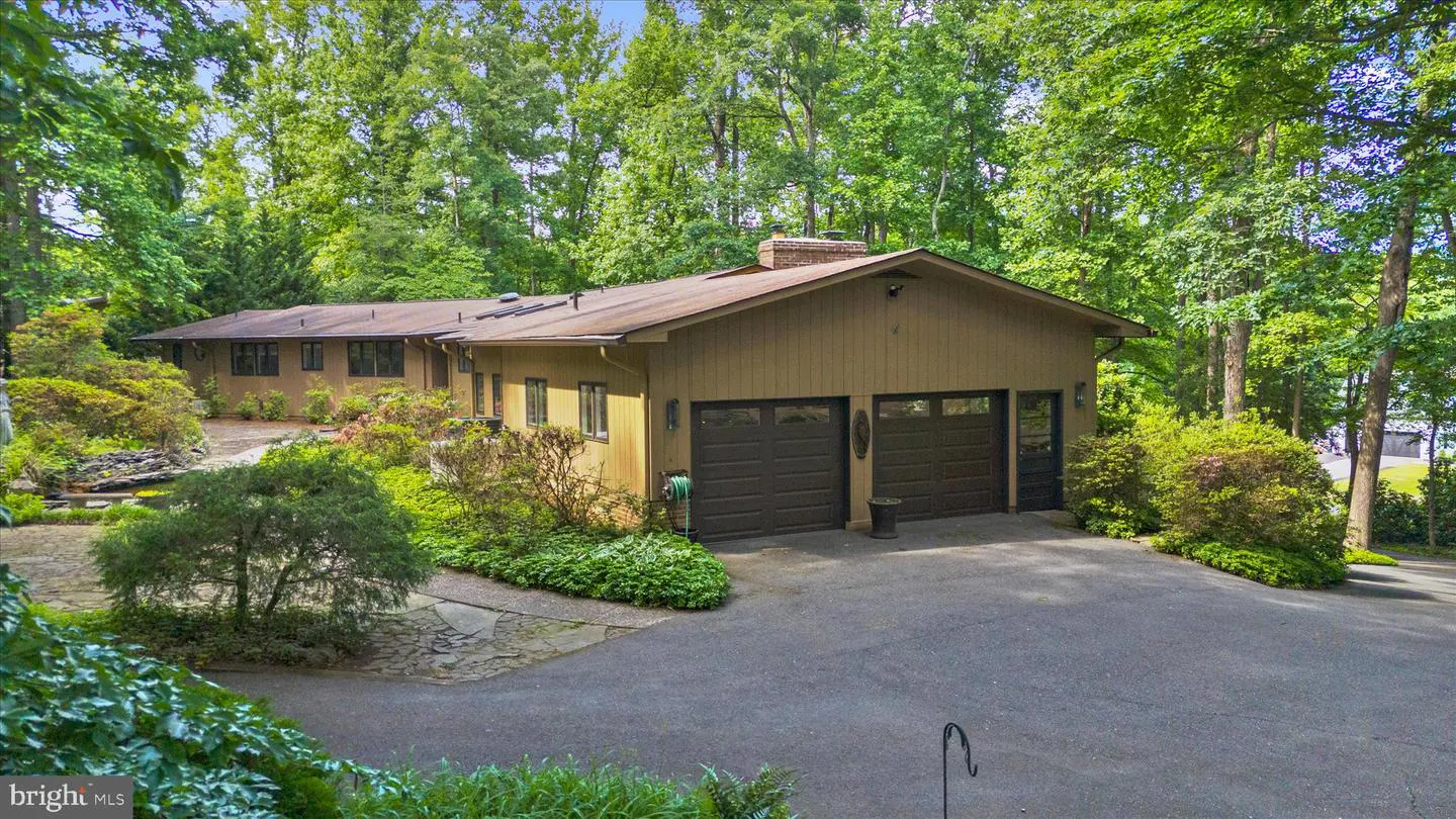 Tan ranch-style home with a two-car garage, paved driveway, and lush green trees in a wooded backyard setting.