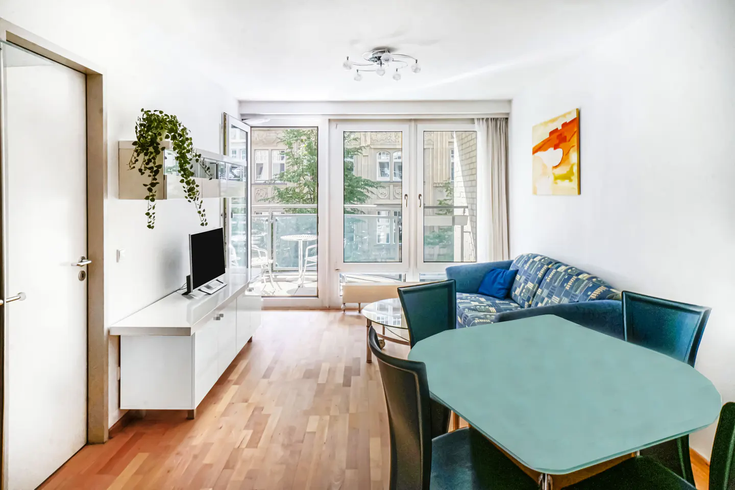 Bright living room with wood floors, white walls, and a balcony. A blue sofa and a light blue table with chairs are visible.