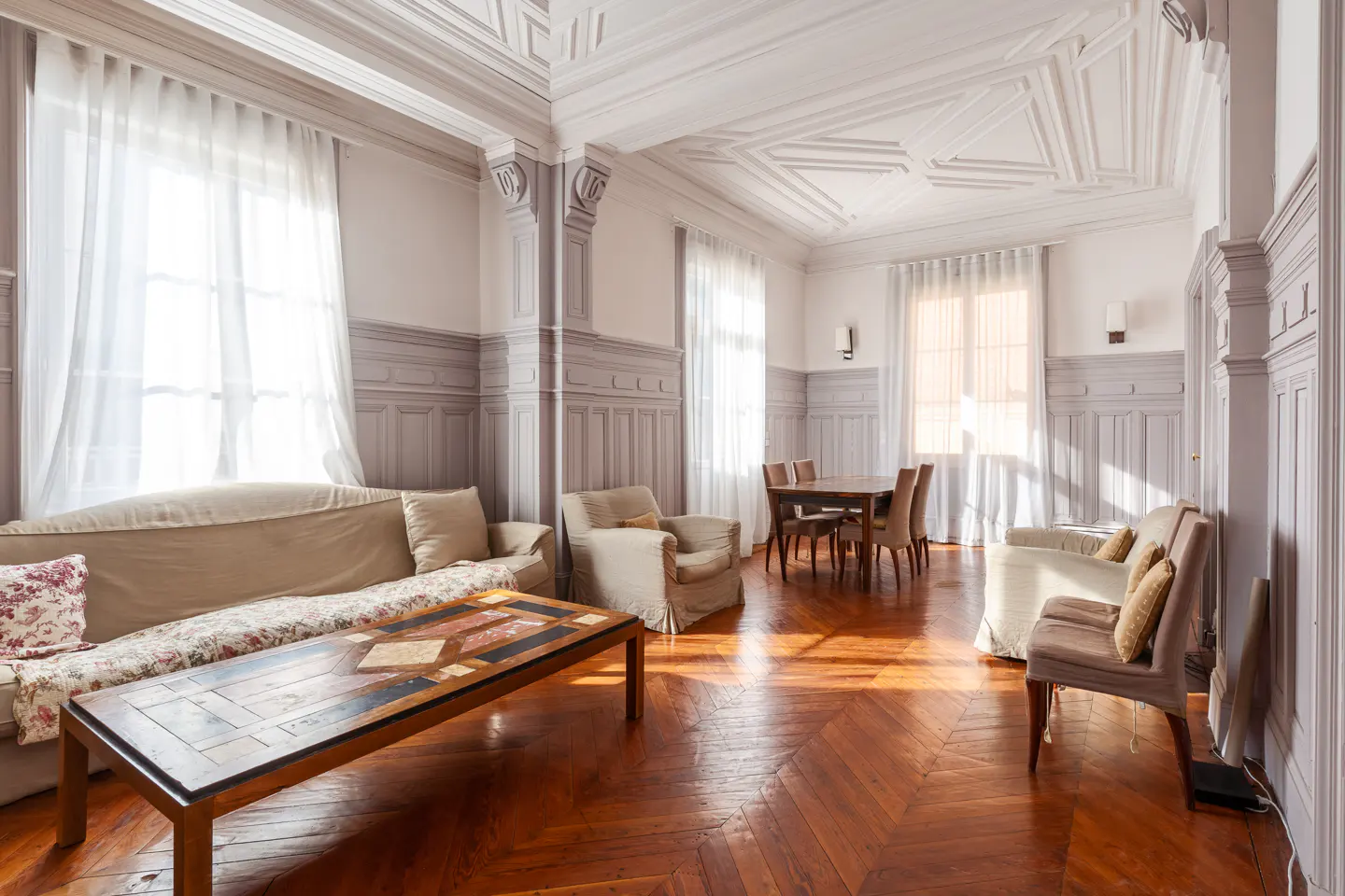 A bright living room with a sofa, armchairs, and a wooden table on a herringbone floor. The walls are paneled in gray, and the ceiling has decorative molding.