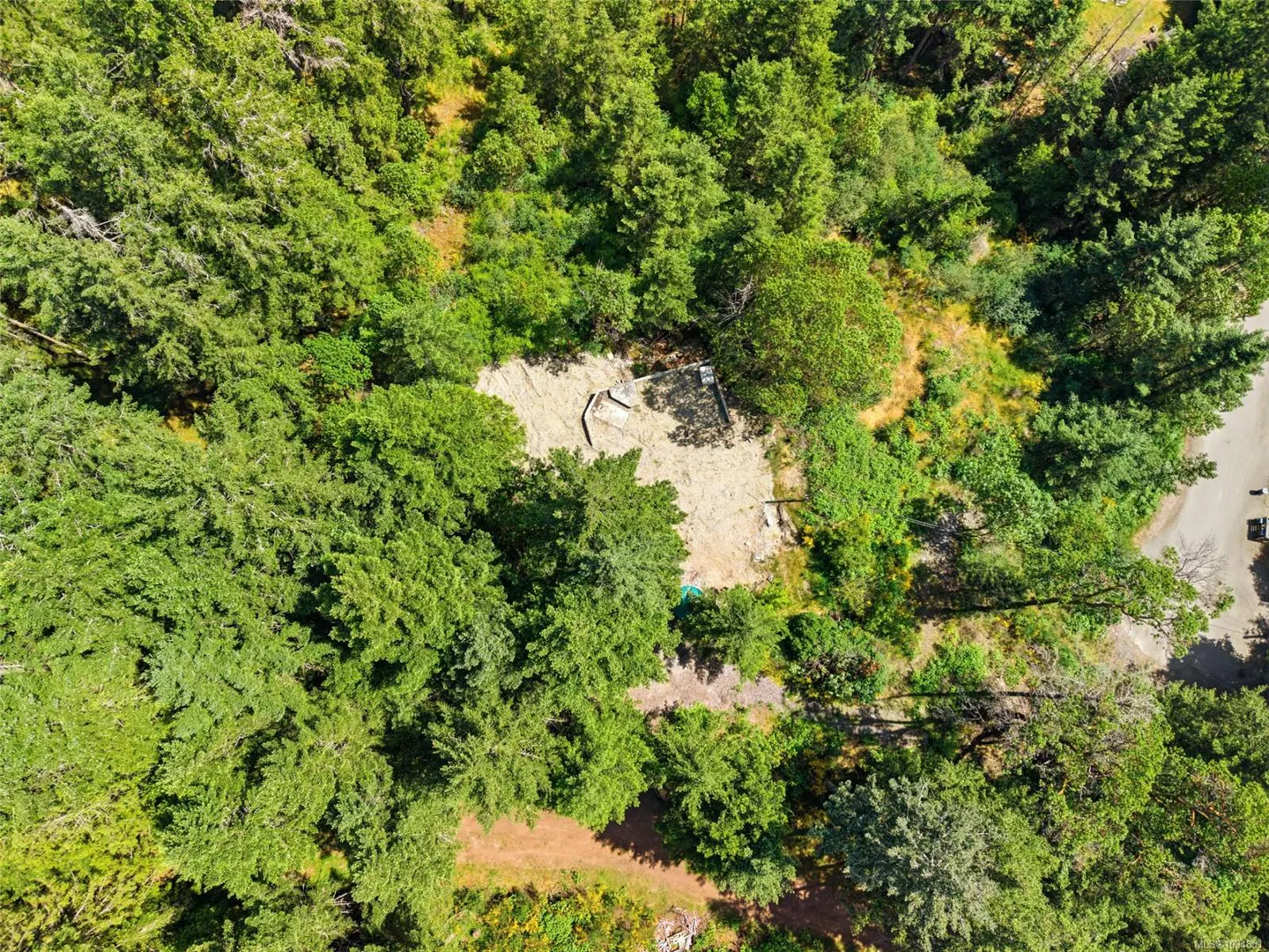 Aerial view of a cleared building site surrounded by dense green forest, with a paved road visible on the right.