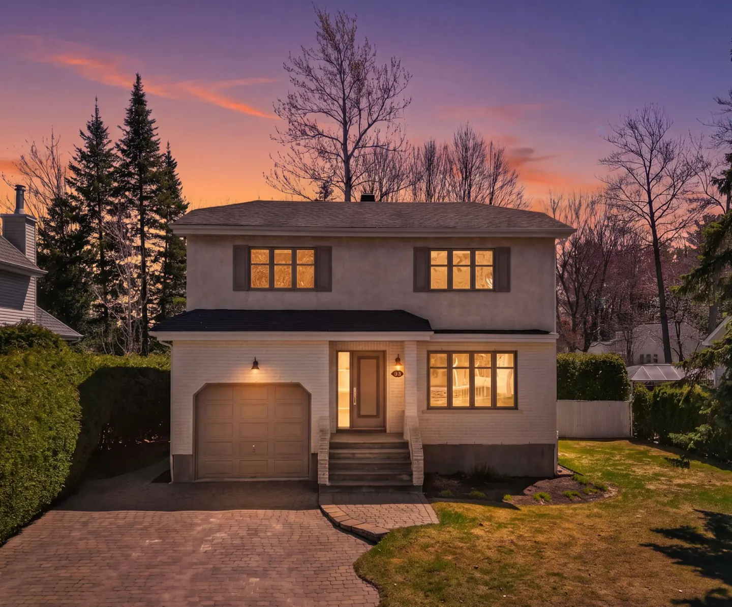 Two-story house with a garage and brick driveway at dusk. The sky is orange and purple.