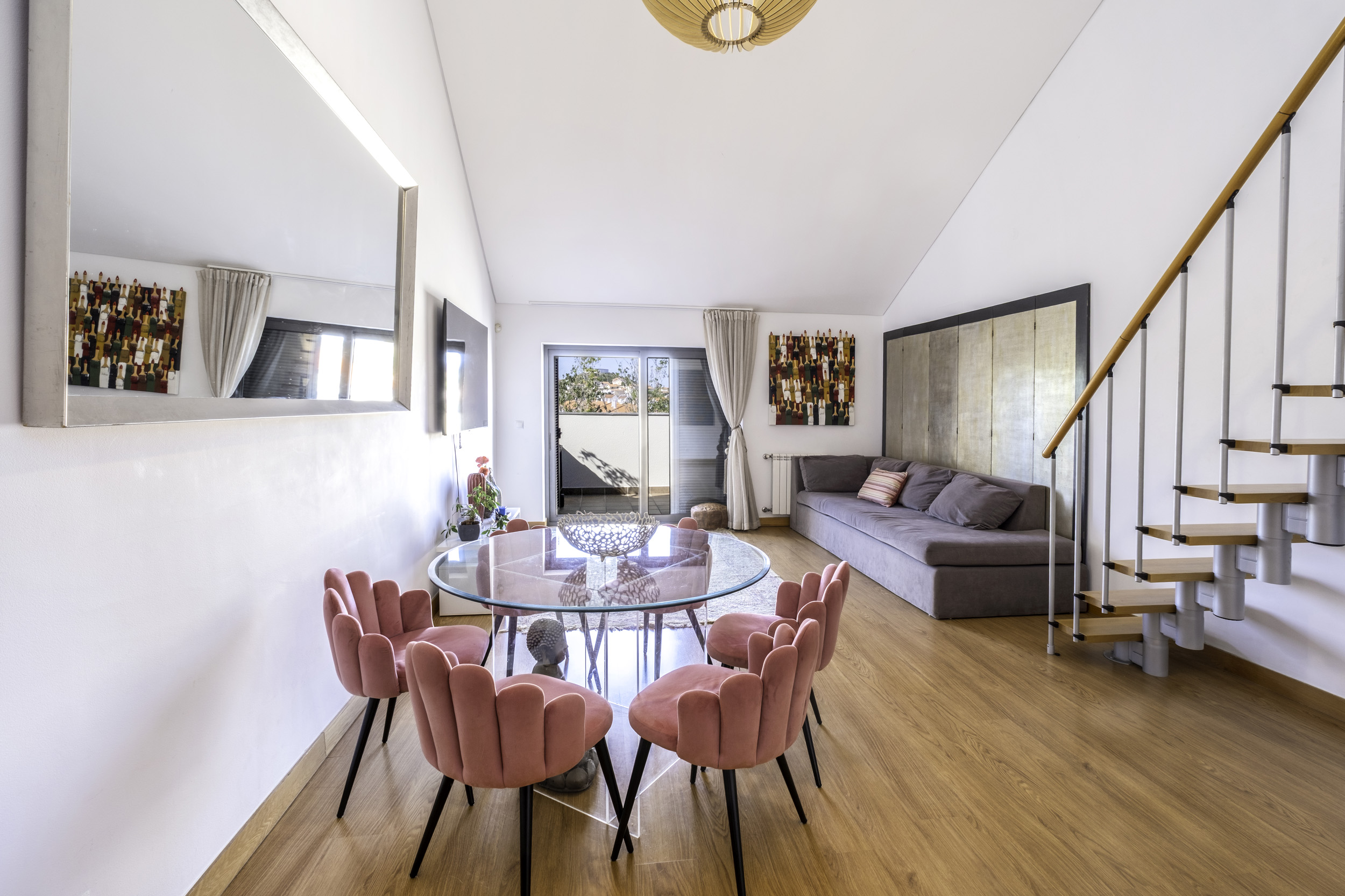 Modern living room with a glass dining table, pink scalloped chairs, a grey sofa, and a staircase on hardwood floors. [AI-generated text]