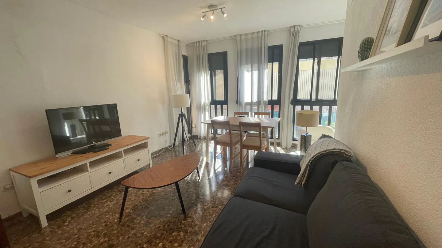 Bright living room with a black sofa, wood table, TV, and dining set near large windows with white curtains.
