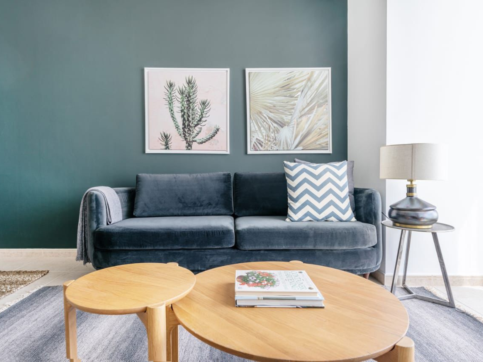 A blue velvet sofa sits against a dark teal wall with two botanical prints, wooden coffee tables, and a side lamp.