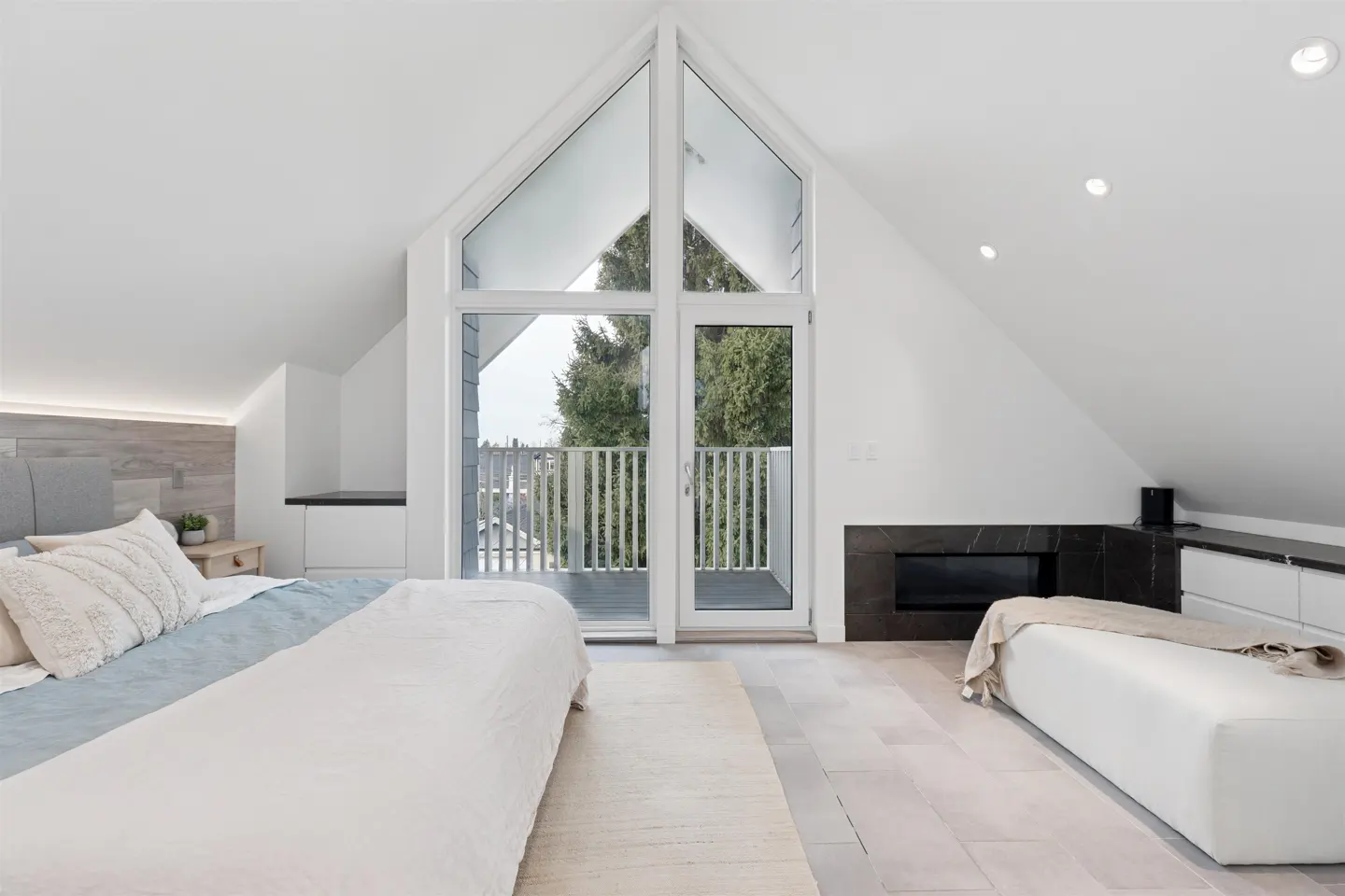 Bright bedroom with white walls, a bed with blue accents, and a balcony with a view of trees. A black fireplace adds contrast.
