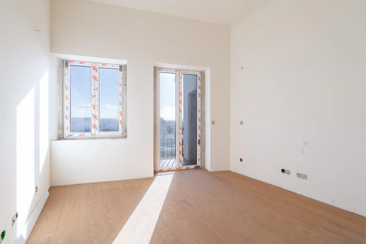 Bright, empty room with light wood floors, white walls, window and glass door to balcony.