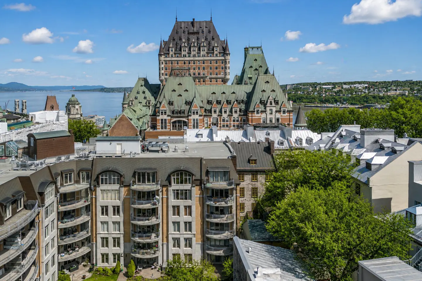 View of Fairmont Le Château Frontenac behind condos with balconies, green trees, and a river in the background under a blue sky.