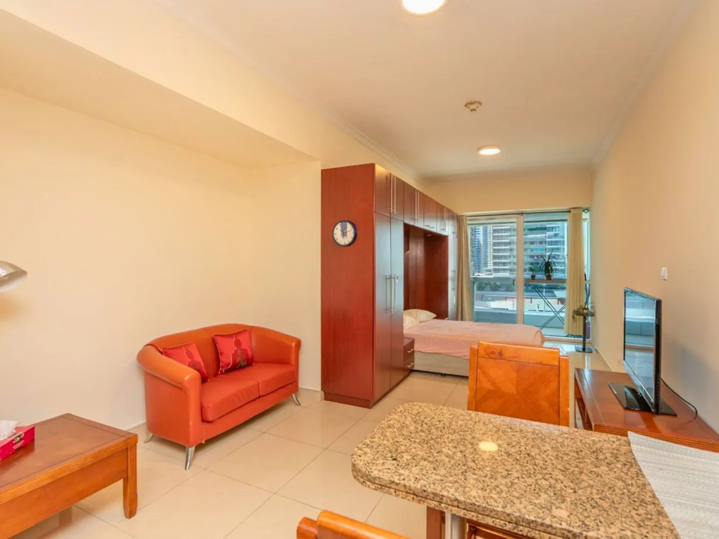 A studio apartment with a red sofa, a granite table, a TV, and a bed in a built-in wardrobe. A balcony overlooks city buildings.