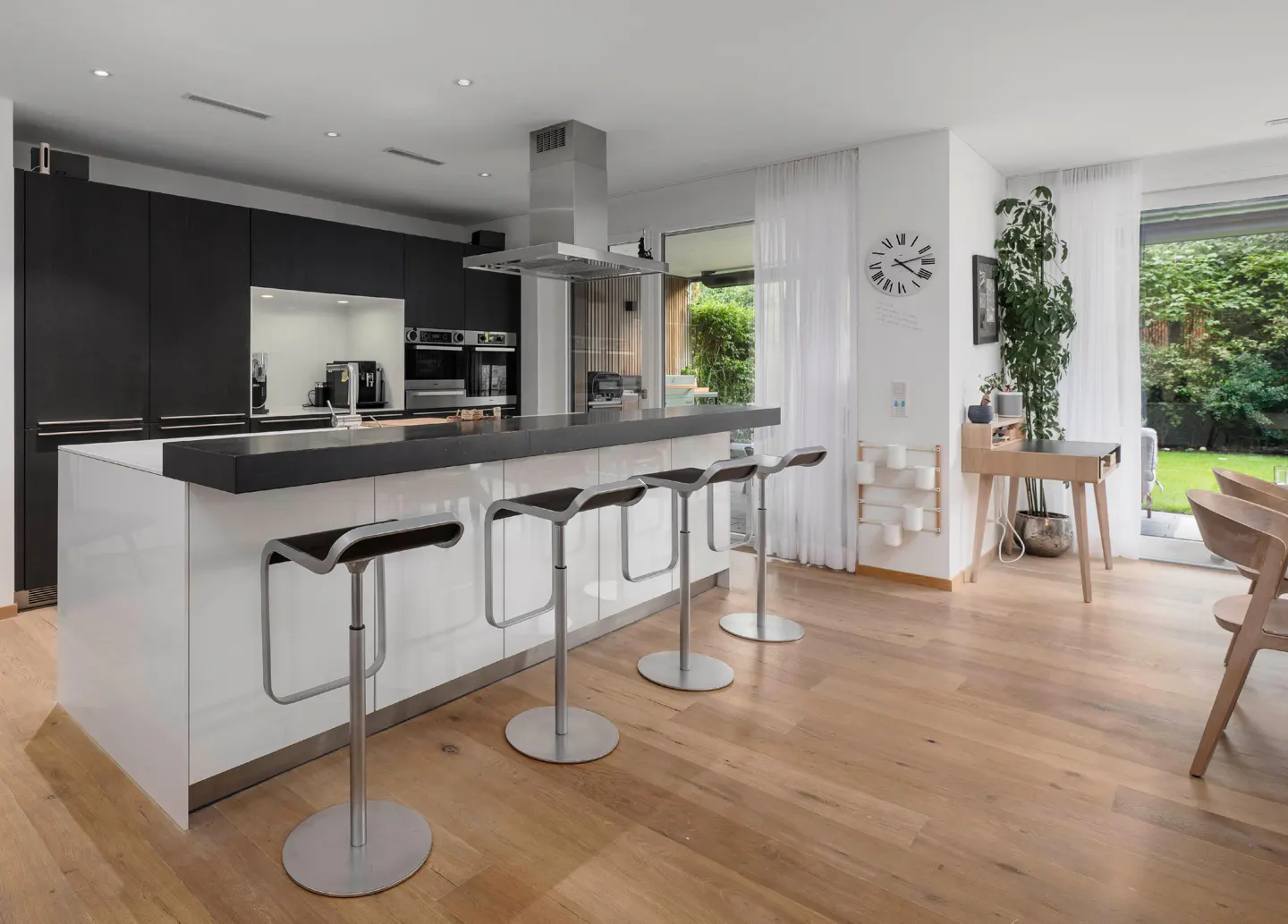 A modern kitchen with black cabinets, a white island with seating, and light wood floors. A large window overlooks a green garden.