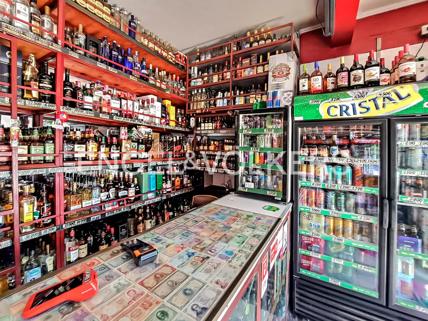 Liquor store interior with shelves stocked with bottles, a counter covered in banknotes, and refrigerators filled with beverages.