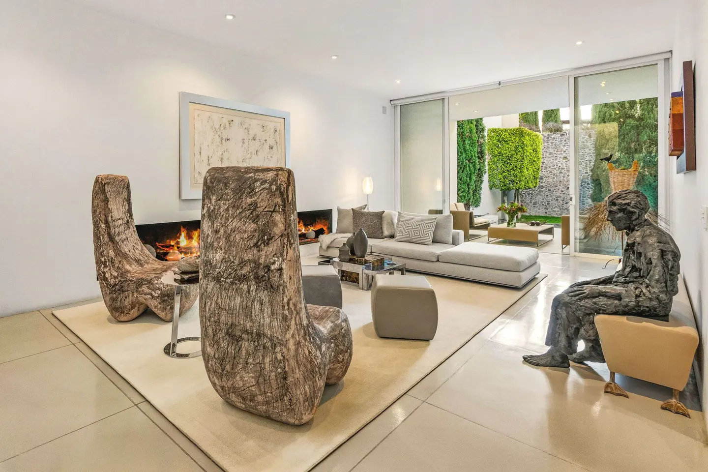 Bright living room with a fireplace, a gray couch, and two wooden chairs. A statue sits near a sliding glass door to a green patio.