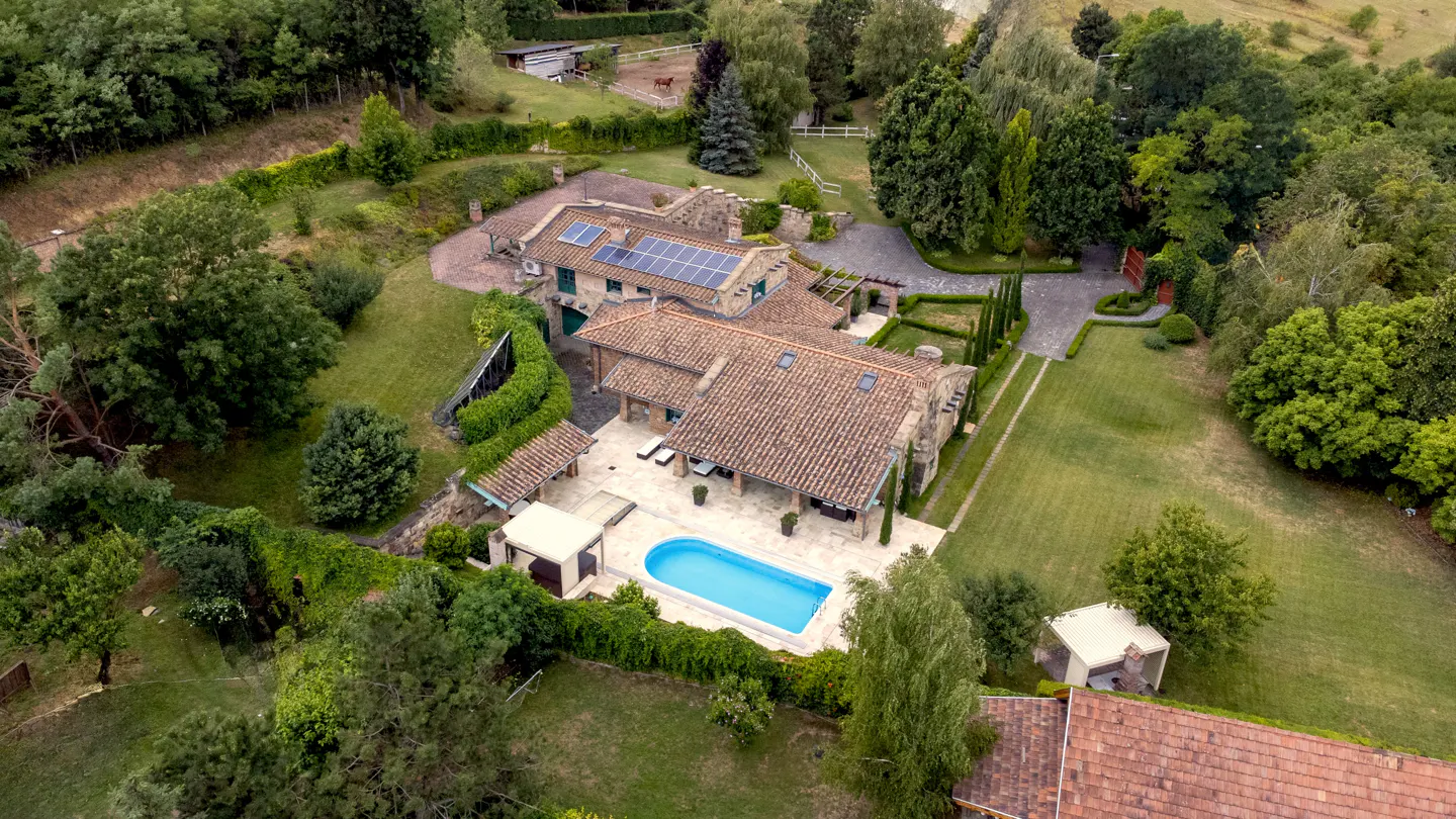 Aerial view of a large stone house with a red tile roof, solar panels, a pool, and lush green landscaping.