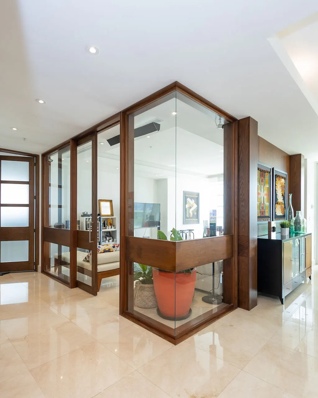 Bright foyer with marble floors, wood-framed glass walls, and a wooden door. A large orange pot with a plant sits in the corner.
