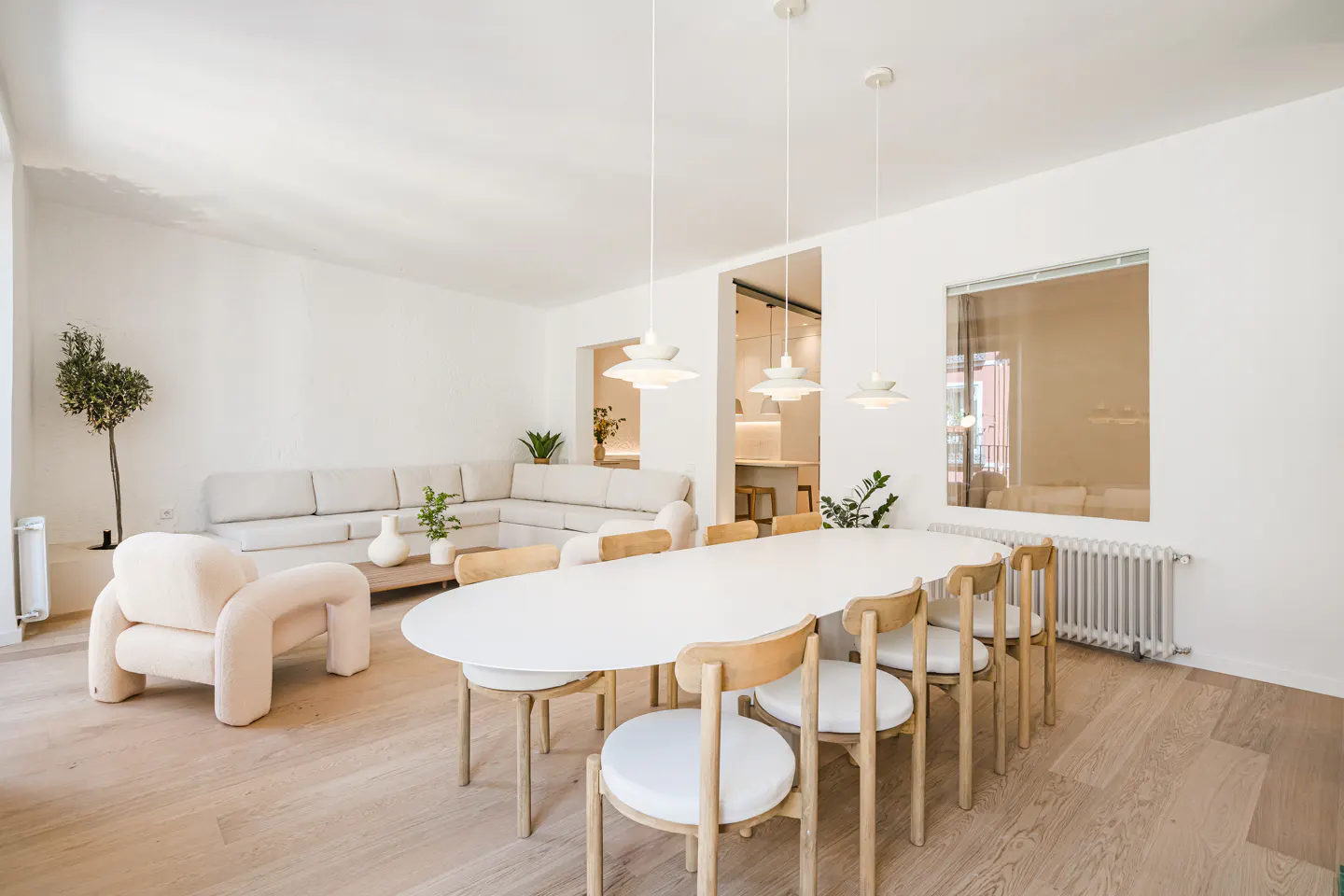 Bright, modern living room with white walls, light wood floors, and minimalist decor. A large white dining table with wooden chairs sits near a beige sofa.