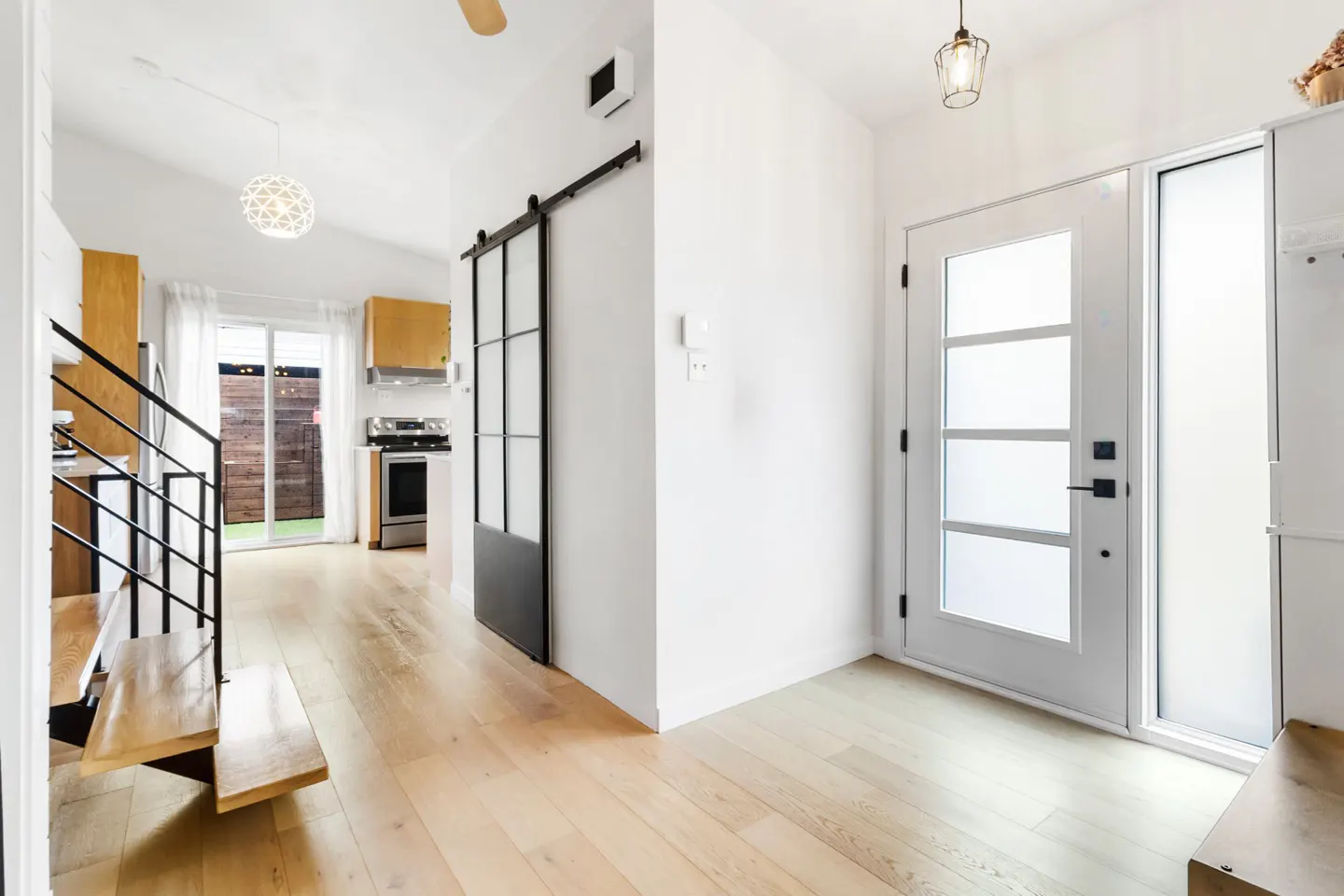 Bright, modern home interior with light wood floors, white walls, and a black sliding barn door. A staircase and kitchen are visible in the background.