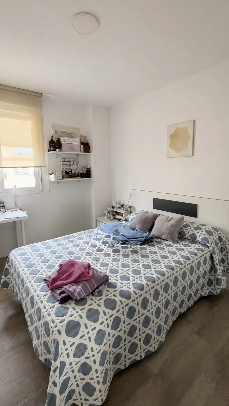 Bedroom with a large bed covered in a blue and white patterned quilt, gray pillows, and pink clothes. White walls, wood floor, and a desk by the window.
