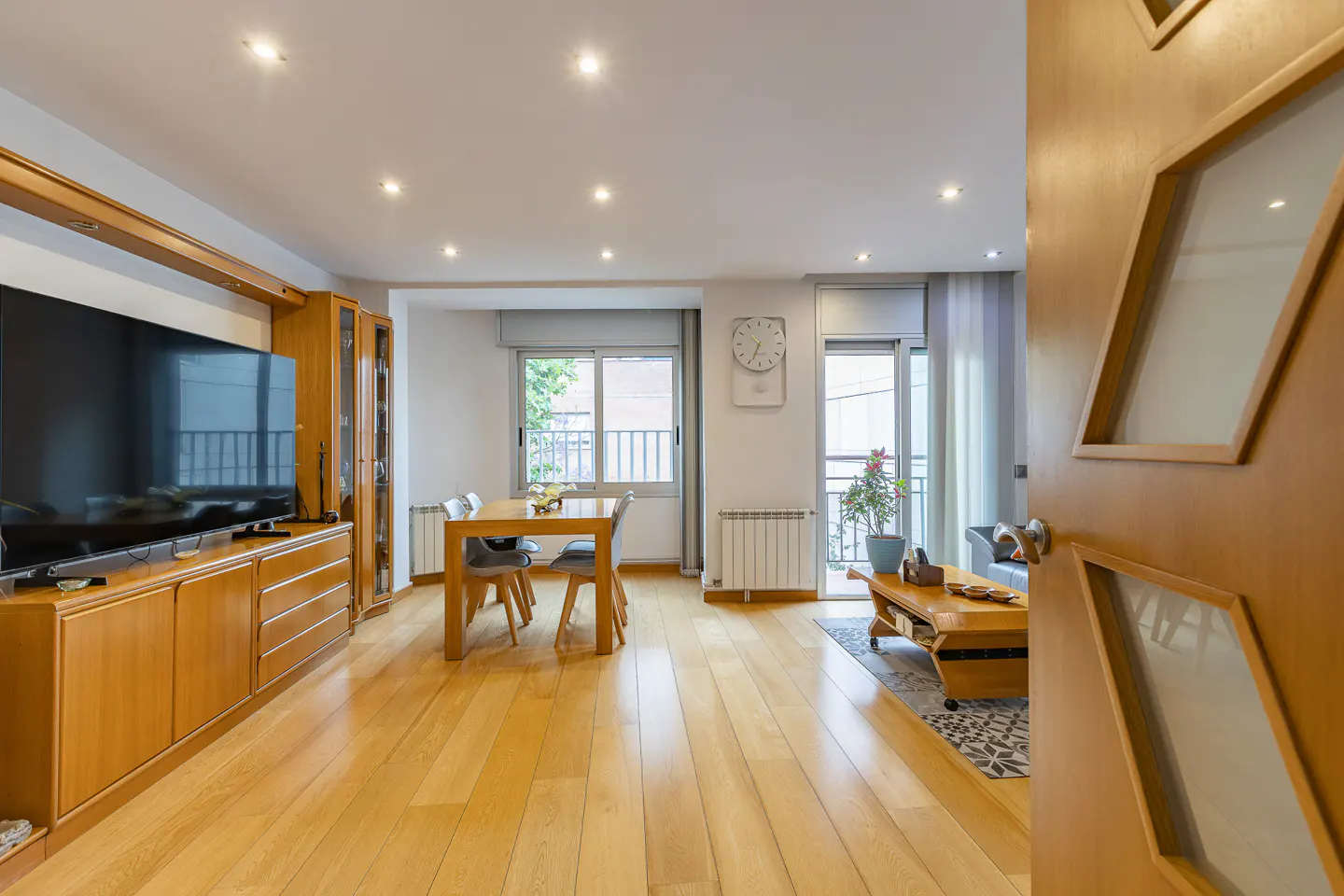 Bright living and dining room with light wood floors, a large TV console, dining table, and a view of a balcony.
