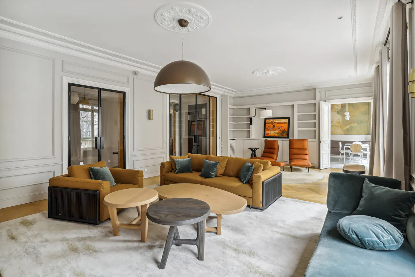 A bright living room with mustard sofas, armchairs, and blue pillows. There are round wooden tables, a white rug, and a large dome pendant light.