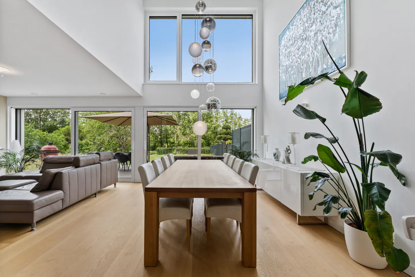 Modern dining room with a long wooden table, white chairs, a hanging glass chandelier, and large windows overlooking a patio.