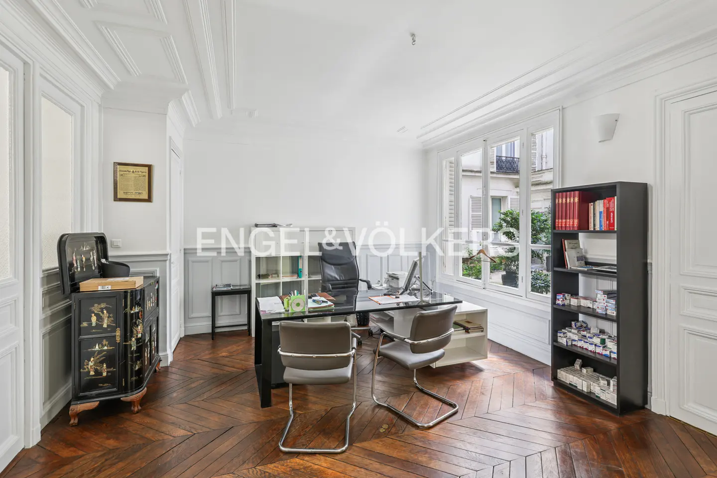 Bright office with herringbone wood floors, white walls, and a black desk with two gray chairs. A black bookcase stands near a window.