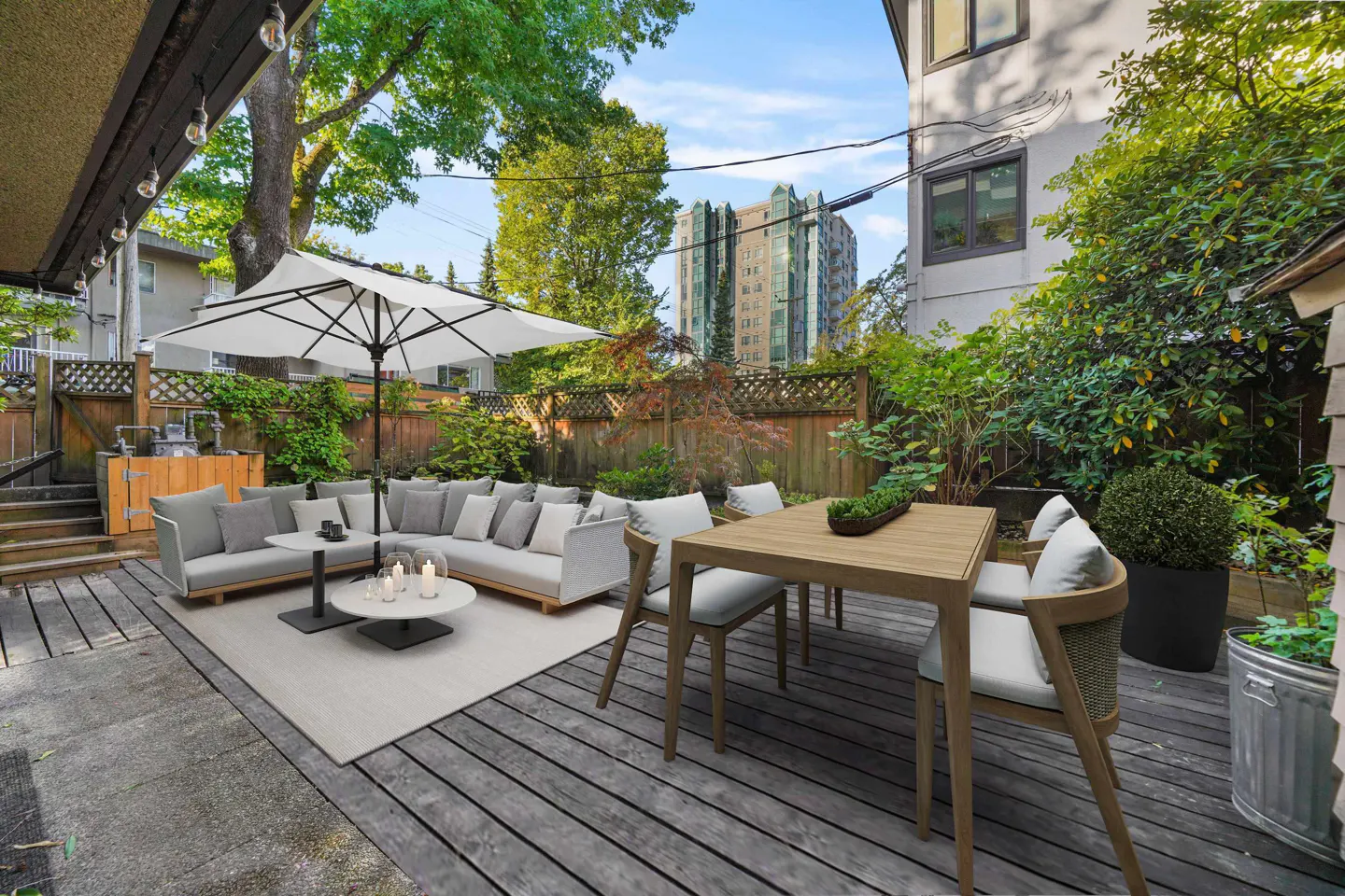 Outdoor patio with gray wood planks, a white sofa set with a white umbrella, and a wooden table with chairs. Green trees and a building in the background.