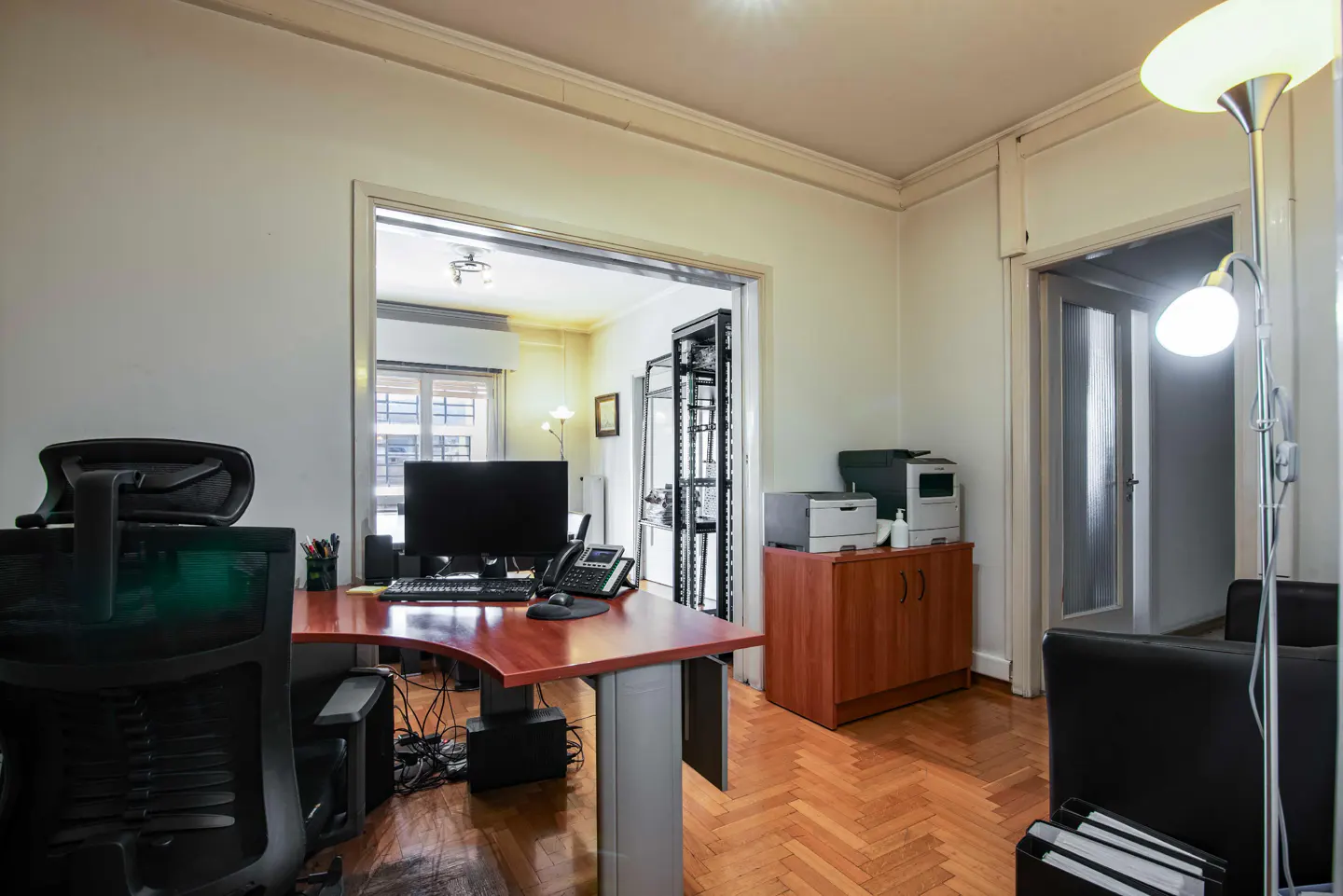 Office space with a wood desk, black chair, computer, and printers on a cabinet. Herringbone wood floors and white walls.
