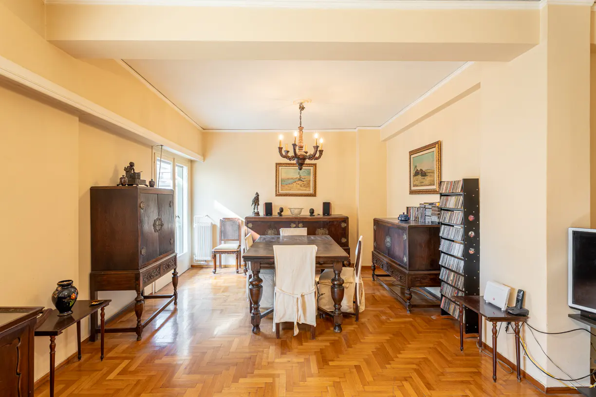 A dining room with parquet floors, yellow walls, and antique furniture. A wooden table with chairs sits under a chandelier.