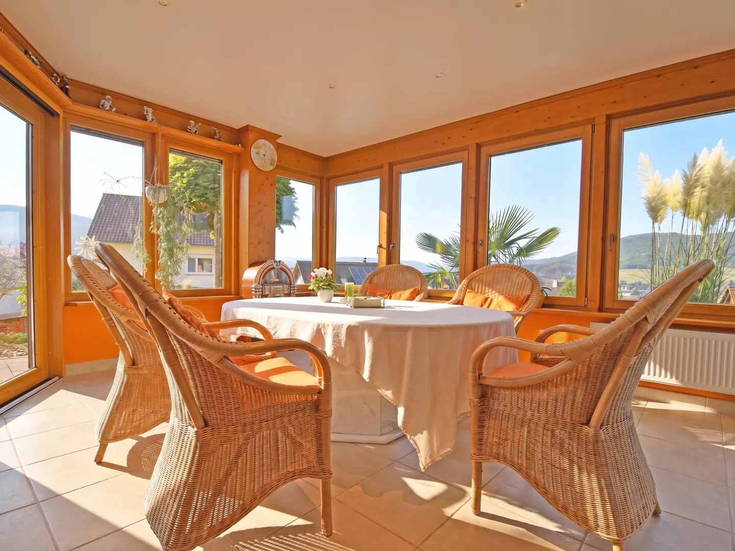 Sunroom with wicker chairs around a white-clothed table. Large windows offer a view of trees and a distant landscape.