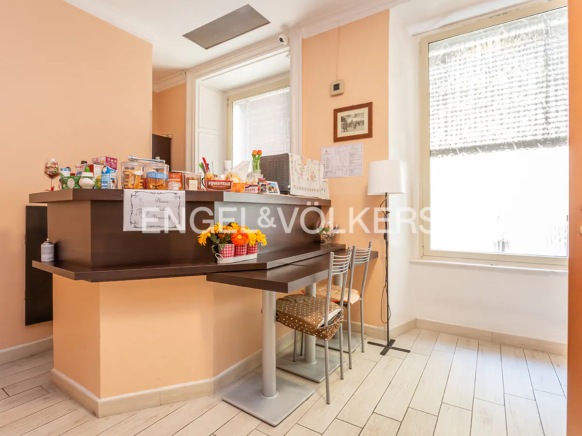 Interior view of a real estate office with a dark wood reception desk, Engel & Völkers logo, and two chairs. A window with blinds is on the right.