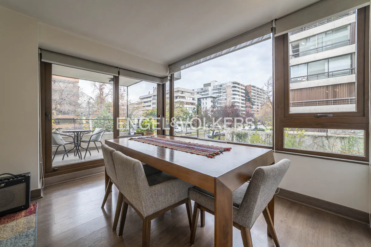 Dining room with a wooden table, grey chairs, and a colorful runner, featuring large windows overlooking city buildings.
