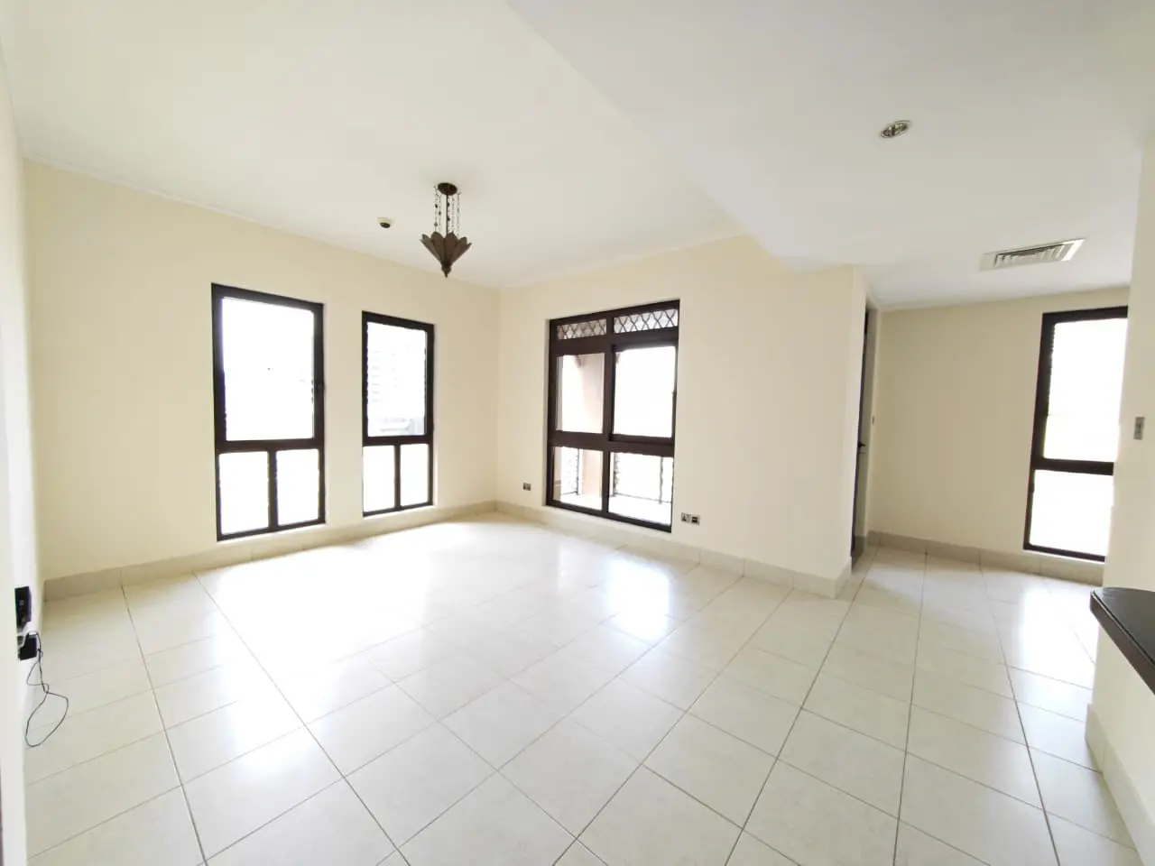 Bright, empty room with white tile floor, cream walls, and dark-framed windows. A chandelier hangs from the ceiling.