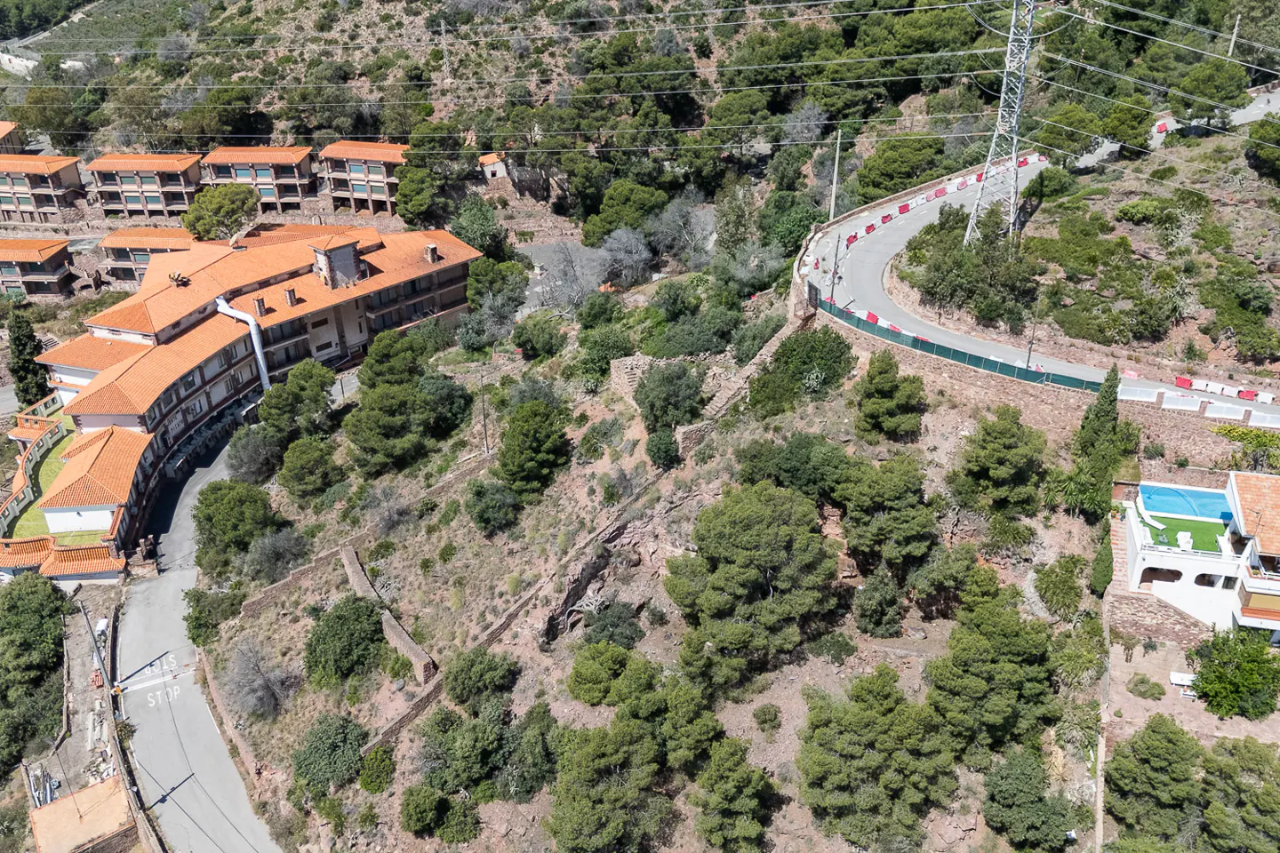 Aerial view of a multi-story building with orange tile roof, surrounded by trees and a winding road on a hillside.
