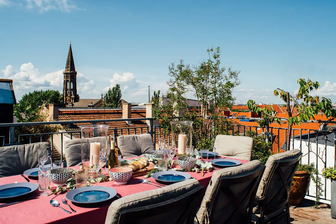 Rooftop dining area with a pink tablecloth, blue plates, and gray cushioned chairs. A church steeple is visible in the background.