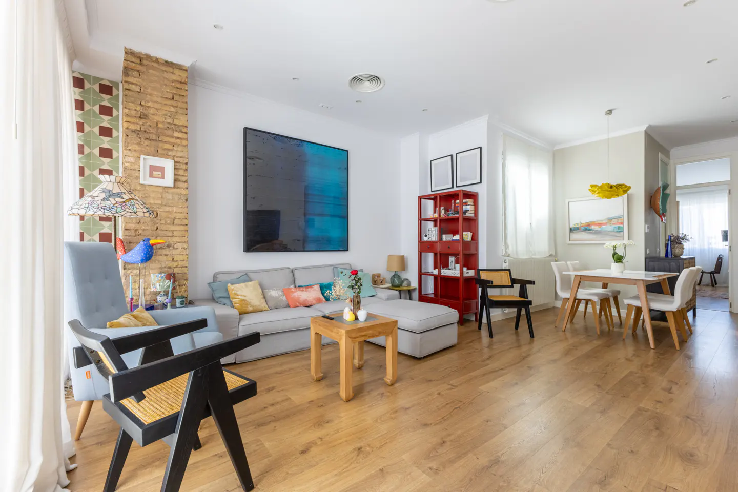 Bright, open-concept living and dining area with wood floors. A gray sofa, blue chairs, and a red bookcase add pops of color.