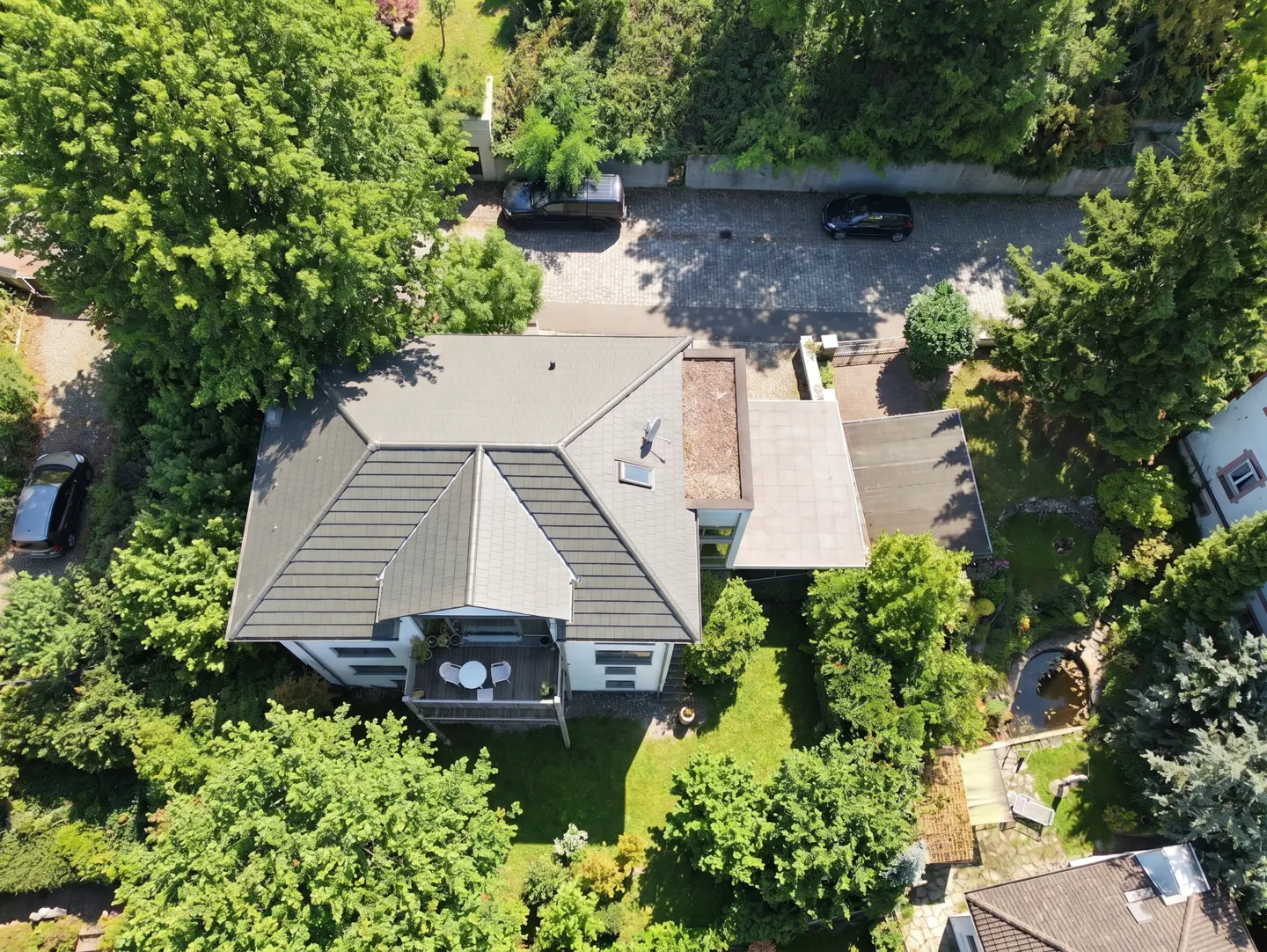 Aerial view of a house with a dark tiled roof, a balcony with patio furniture, and a lush green garden with a small pond.
