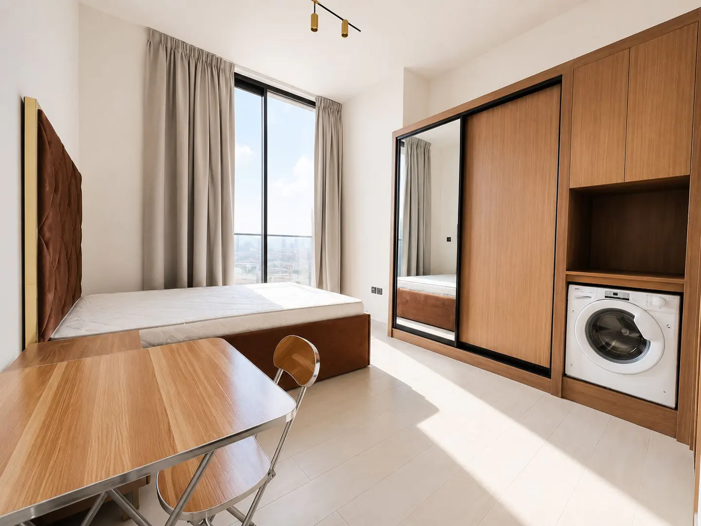 Studio apartment with a bed, table, wardrobe with sliding doors, and a washing machine. Large window with beige curtains.