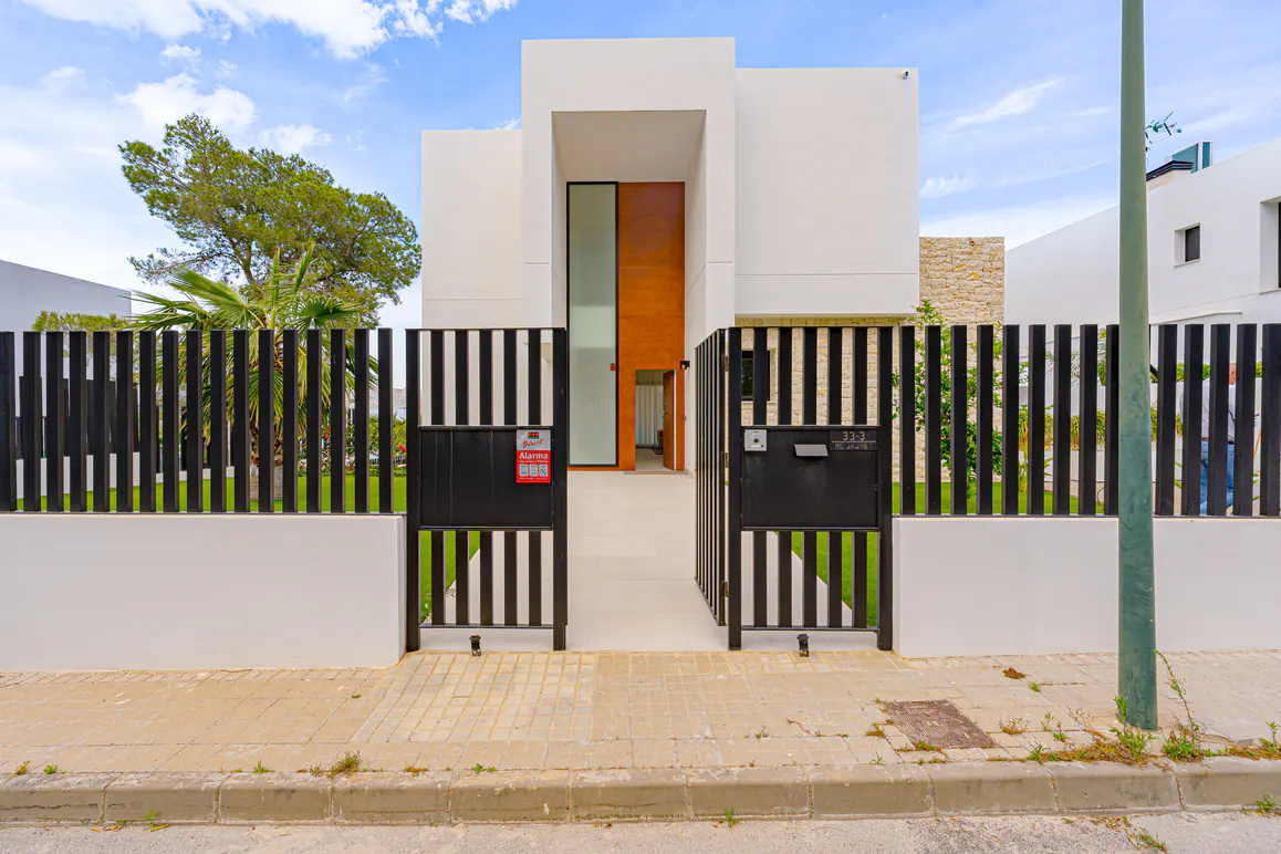 Modern white house with a black striped gate open to the front door. A green tree is visible behind the fence.