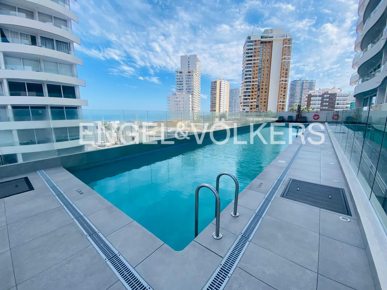Rooftop pool with metal ladder and grey tile surround. Ocean and city buildings are visible beyond a glass barrier. Blue sky.